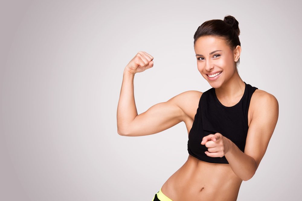 mladá usmiata žena s napnutými svalmi bicepsu na šedom pozadí